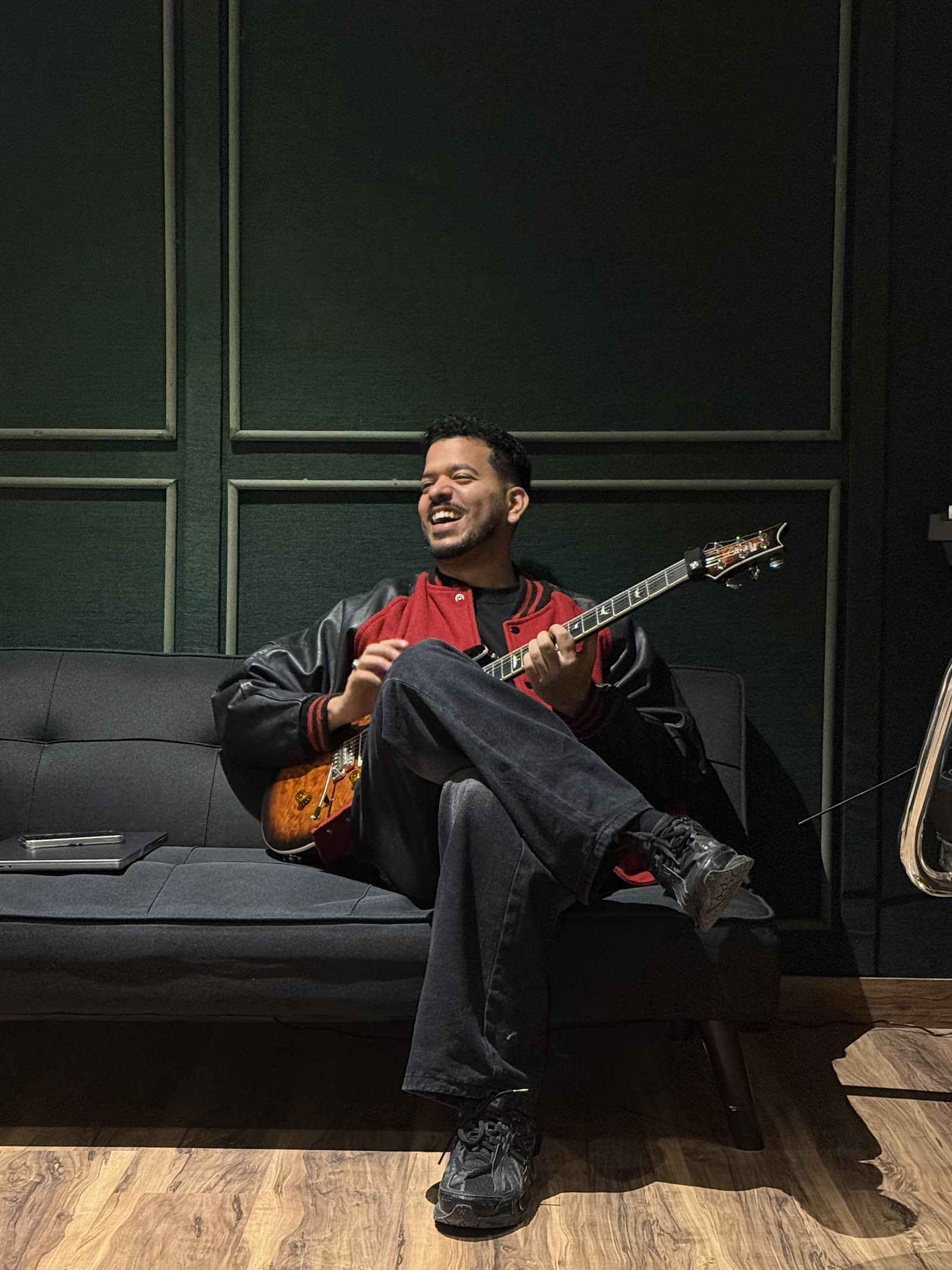 Semwal laughing with a guitar on a couch against a green-panelled wall